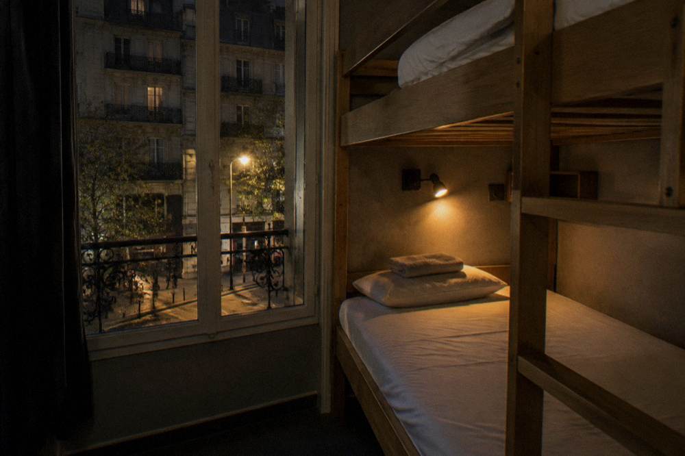 A single hostel bunk with a made bed, a reading light and a folded towel on the pillow