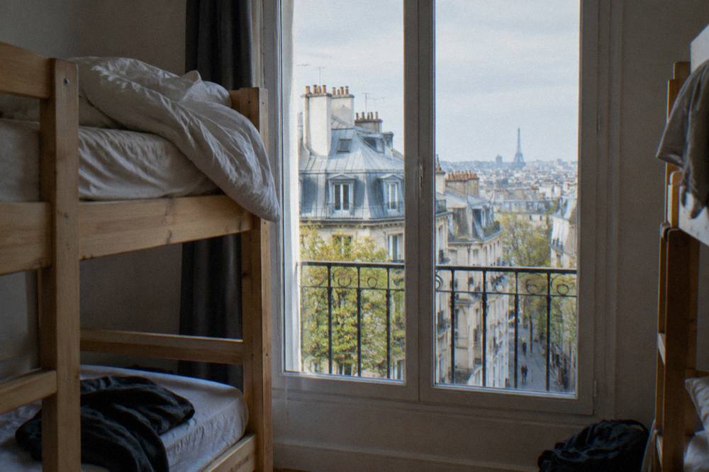 Bunk beds in a bright hostel dorm room with a window onto Paris rooftops