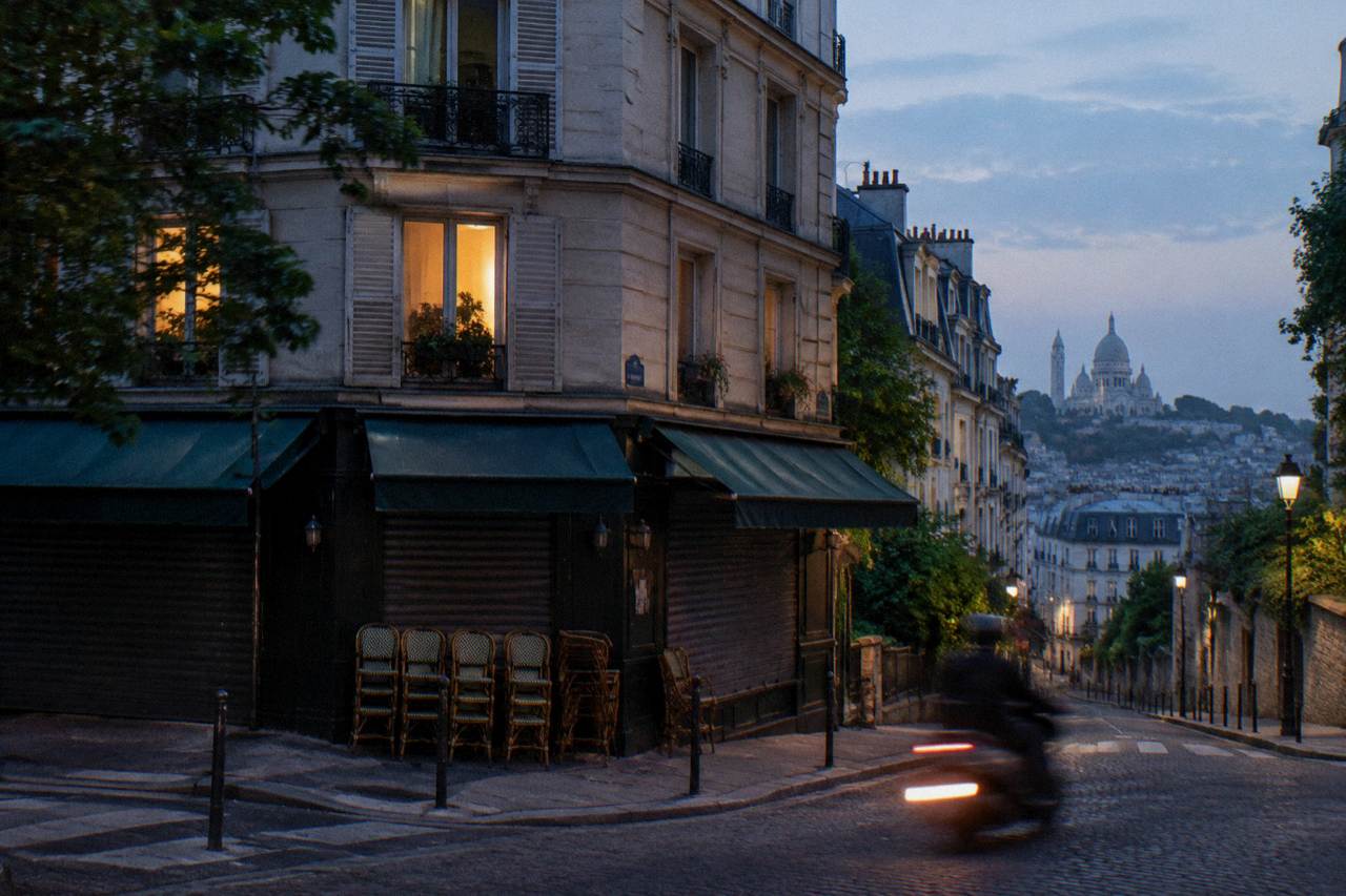 A quiet Paris street at dusk with hotel windows lit above a shuttered cafe