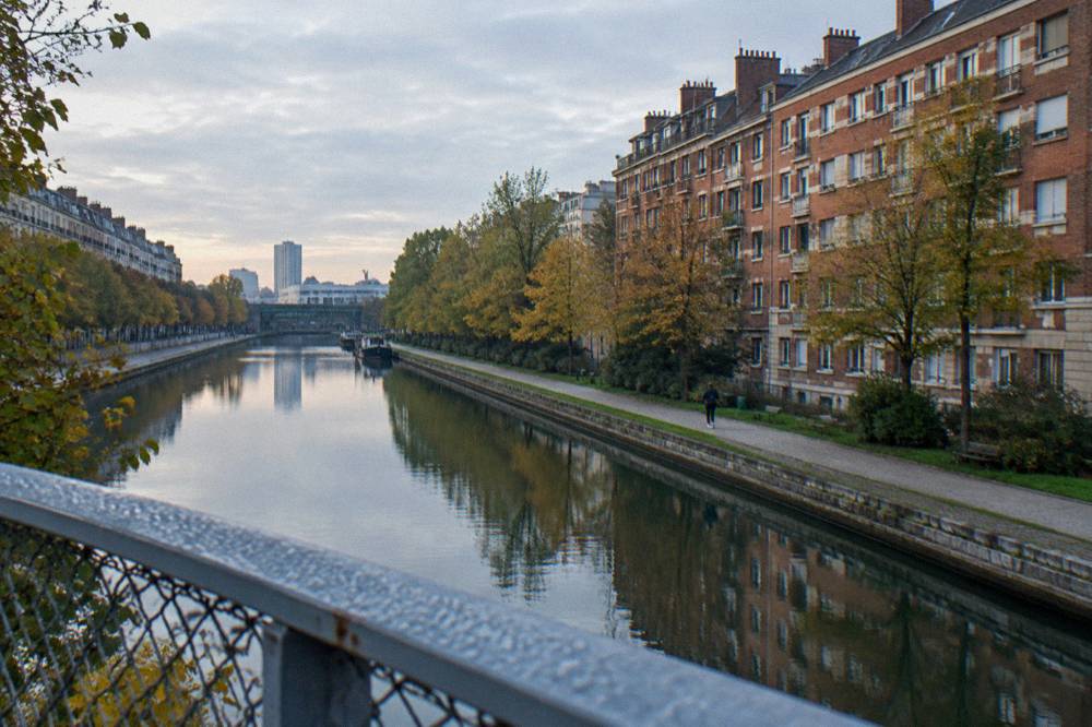A canal-side street in northern Paris with modern residential buildings and a footbridge