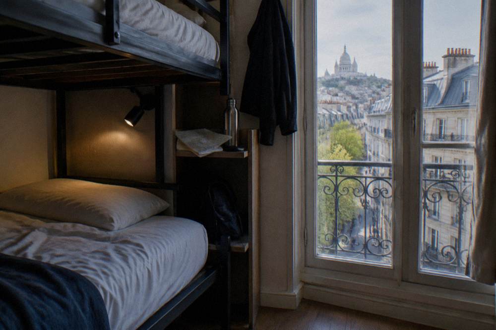 A single hostel dorm bed with a reading light and personal shelf beside a tall window
