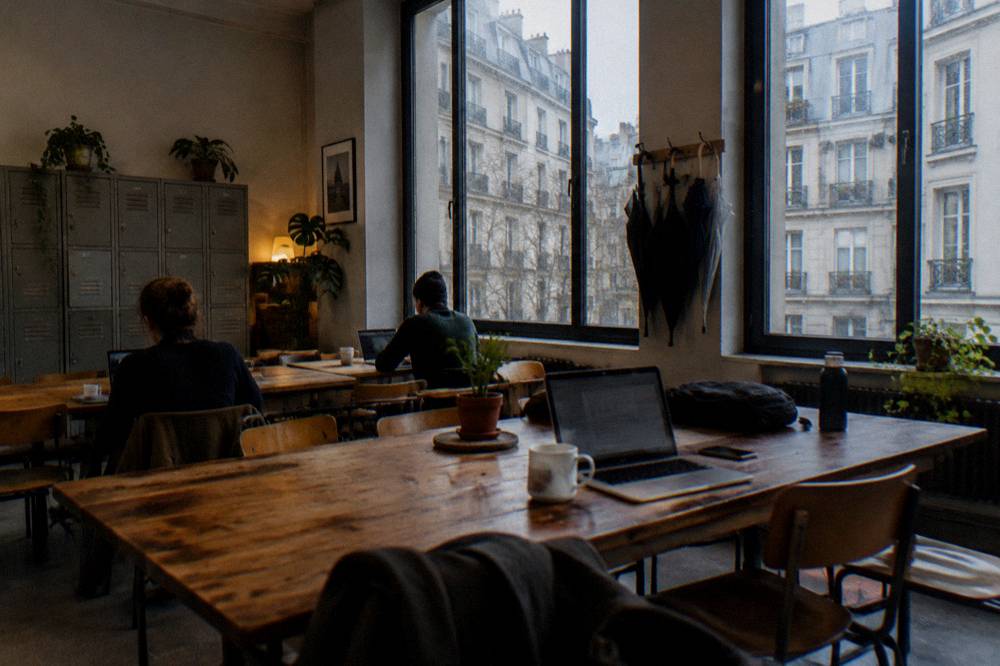A hostel common room with wooden tables, lockers along the wall and large windows onto a Paris courtyard
