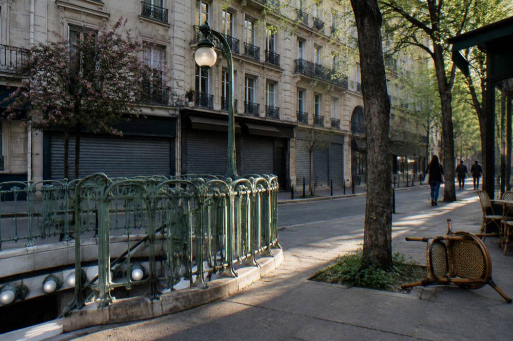 A Paris metro entrance sign on a quiet morning street with shuttered shopfronts below stone apartment buildings